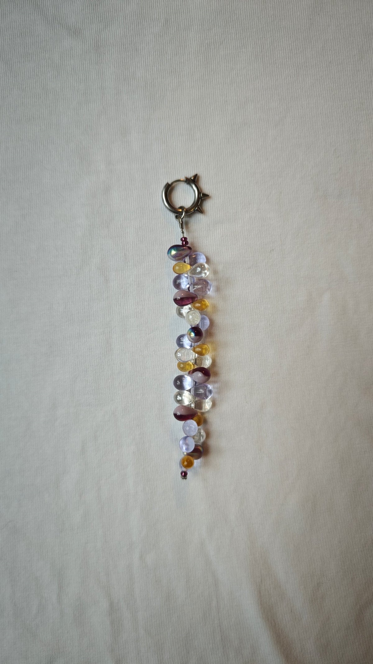 Mono boucle d'oreille de gouttes de verre lilas, ambre, transparentes et violet irisé, avec un anneau à pics en acier, posée à plat