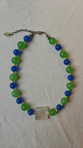 Collier de perles de verre bleues et vertes avec un cube transparent aux reflets argentés au centre, posé à plat
